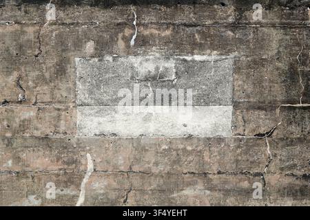 A weathered concrete wall with visible cracks and discoloration. The surface is rough, with rectangular patch of lighter gray is in the center. Stock Photo