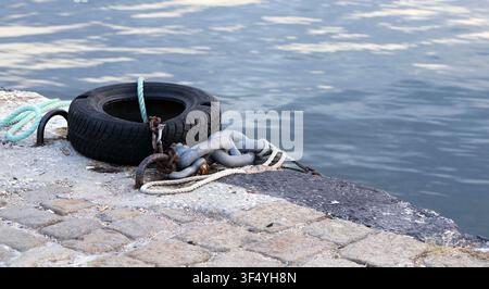 Used tire fender, heavy metal chain, and mooring ropes resting on a weathered stone paved pier, showcasing nautical equipment by the calm ripples of w Stock Photo
