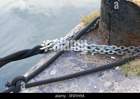 Naval mooring bollard providing a secure anchor point on a dock, holding a thick black rope and a heavy metal chain, symbolizing strength and connecti Stock Photo