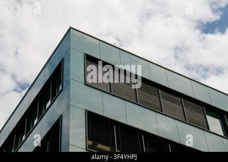 Low angle horizontal view of a modern corporate office building corner with green-gray metallic cladding and dark glass windows against blue sky Stock Photo