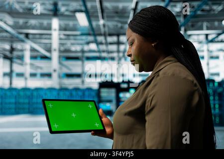 Data center admin monitors components for potential failures using green screen tablet, improving system stability. Server hub worker uses chroma key device to configure settings, increases uptime Stock Photo