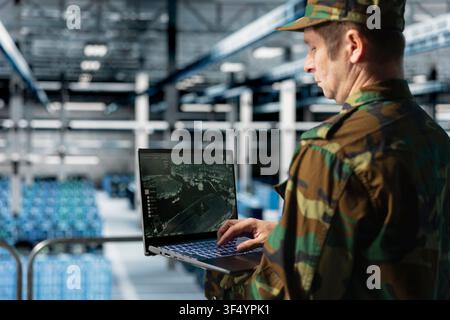 Army specialist in data center uses aerial image overlay on laptop to confirm enemy target locations. Military man in server room performing wide-area scan for threat detection using satellite systems Stock Photo