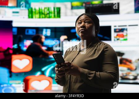 Woman browses social media feed on smartphone in front of digital video wall showcasing multimedia content. Viewer using phone in modern space surrounded by diverse multimedia clips Stock Photo