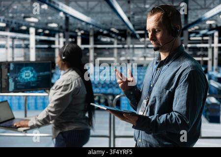 Team of data center colleagues using headset mic, interpreting AI logs to resolve clients issues. Technicians identifying hardware issues using predictive artificial intelligence alerts, guiding users Stock Photo