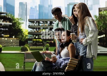 International students gather around laptop on campus bench, reviewing coursework notes and ideas together during outdoor learning collaboration. Group study project discussion. Stock Photo