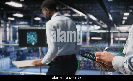 Close up of server room worker checking code on tablet next to colleague reviewing AI LLM visualization. Data center IT expert using device to monitor artificial intelligence systems, Stock Photo