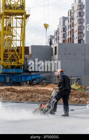Construction worker in orange helmet cutting concrete slab with dust cloud at building site, tower crane and residential buildings under construction Stock Photo