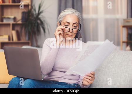 Senior Woman Reading Documents At Home Office With Laptop, Focused Attention, Business Concept Stock Photo