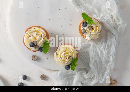 Three cupcakes topped with piped white frosting, fresh blueberries, mint leaves, and chia seeds are arranged on a white marble board with a soft textu Stock Photo