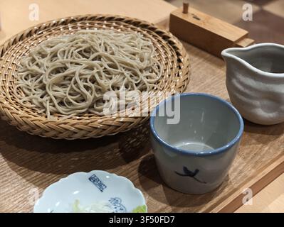 Wheat noodles rest in a woven basket. Simple tea set sits beside them on wood. Calm, minimalist setting feels warm and inviting. Perfect for food, wel Stock Photo