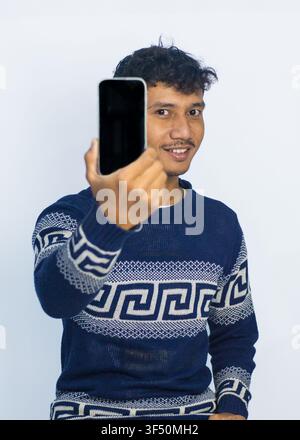 A cheerful young man wearing a patterned blue sweater, smiling while holding and pointing at a smartphone with a blank screen. Studio portrait against Stock Photo