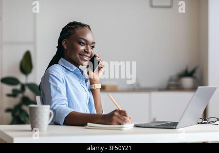Millennial Black Woman Manager Taking Notes And Talking On Cellphone In Office Stock Photo