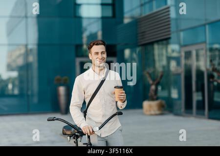 Coffee takeaway and eco transport. Smiling man holding glass and go with bicycle near office building Stock Photo