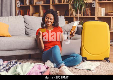 Smiling Black Woman Shopping Online With Laptop And Credit Card At Home, Preparing For Trip Stock Photo