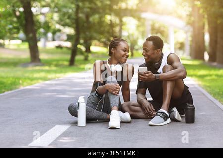 Cheerful Black Athlete Couple Relaxing On Path In Park After Training Outdoors Stock Photo