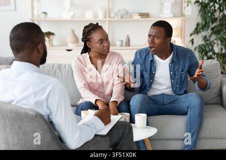 Young Black Spouses Having Relationship Problems, Sitting At Therapy Session With Counselor Stock Photo