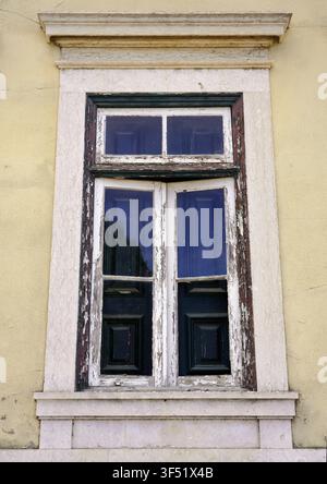A close-up view highlights a weathered wooden window with peeling paint and a stone frame set into a yellow building facade in Lisbon, Portugal Stock Photo