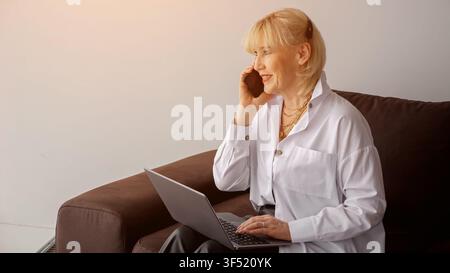 Mature blonde woman managing tasks from home using a laptop and conversing on a smartphone, representing remote work, communication, and connectivity Stock Photo