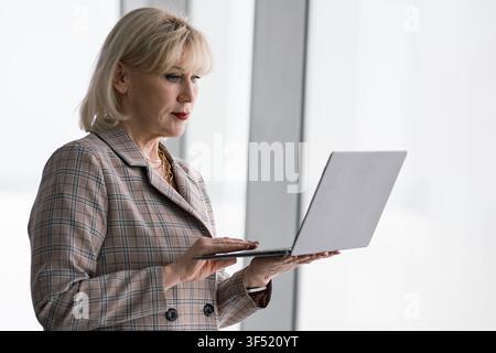 Experienced businesswoman working on a portable laptop, standing in a brightly lit corporate office environment, concentrating on her online tasks and Stock Photo