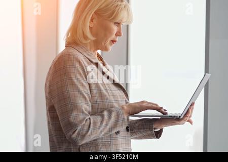 Senior business woman standing and focusing on her laptop, actively engaged in work, representing professionalism, experience, and dedication in a Stock Photo