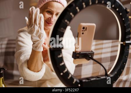 Senior woman wearing moisturizing gloves during evening hand skincare routine while recording beauty blog at home. Anti-aging care and self-care Stock Photo