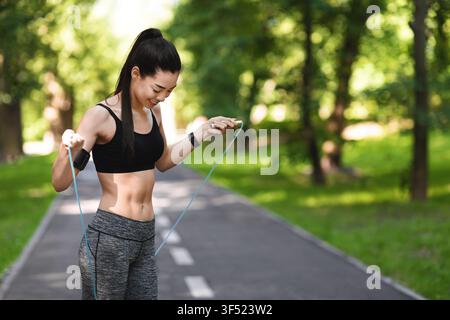 Exercising Outdoor. Fit Young Asian Woman Training With Skipping Rope In Park Stock Photo