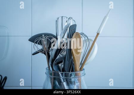 An assortment of kitchen utensils whisks, tongs, a spatula, and a wooden spoon stands upright in a glass jar against a white tiled kitchen wall. Stock Photo
