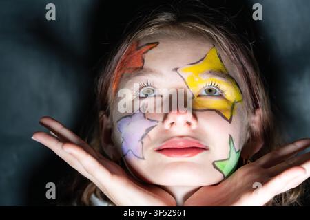 Close-up portrait of a preteen girl with colorful star face painting looking upward and posing with hands under her chin against a dark background. Stock Photo