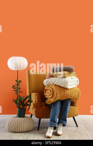 Woman with warm plaids sitting in armchair near orange wall in room Stock Photo