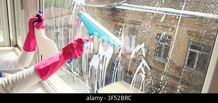 Person in pink gloves cleaning a window with a squeegee and spray bottle, household chores and spring cleaning concept, the focus is on the hands and Stock Photo