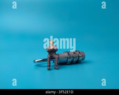A miniature businessman sitting on top of a silver combination padlock against a plain blue background. Stock Photo