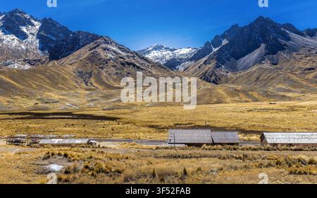 Majestic Apu Chimboya rising above the high Andean plateau with snow‑capped peaks, golden grasslands and rural mountain shelters under a clear blue sk Stock Photo