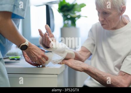 Nurse helping with bandage on elderly man in clinic. Healthcare provider assisting senior male patient with compassion and gentleness. Aging man support and elderly care in medical facility room Stock Photo