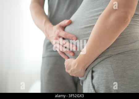 Pregnant woman alone cradling belly with interlaced fingers in serene solo portrait. Expecting mother embracing motherhood in minimal studio with white background showing protective anticipation. Stock Photo