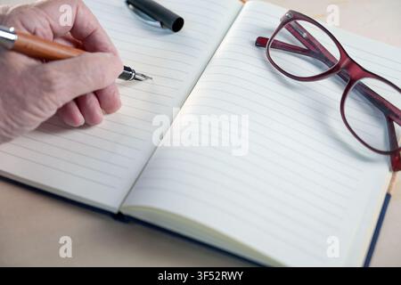 Older hand with a fountain pen writing in a notebook with blank lined paper, glasses laying on the side, concept image for handwriting, diary, notes, Stock Photo