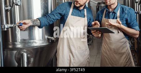 Two Men Working In Brewery, Checking Beer Tanks And Taking Notes, Industrial Setting, Unrecognizable Stock Photo
