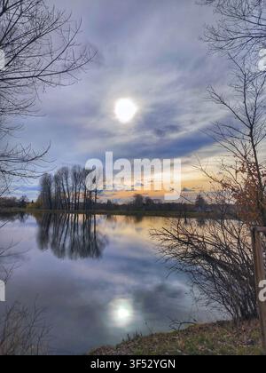 Beautiful scenic landscape of a calm lake reflecting cloudy sunset sky and silhouette of trees with the sun shining through clouds Stock Photo
