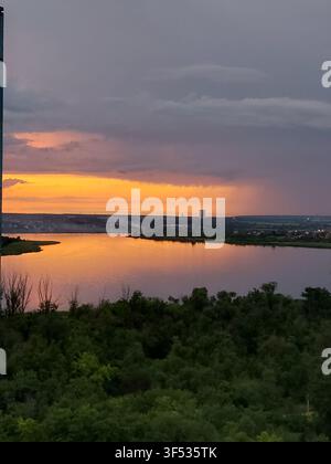 Scenic sunset over a tranquil lake with dramatic storm clouds, distant rainfall, and colorful reflections on the water. Peaceful evening landscape featuring vibrant orange sky, lush green forest, and approaching summer storm. Stock Photo