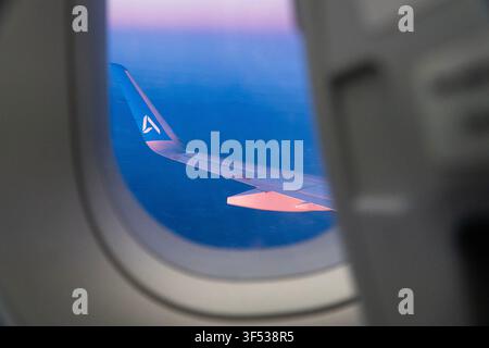 A passenger window shows an airplane wing flying over a blue ocean during twilight. Stock Photo