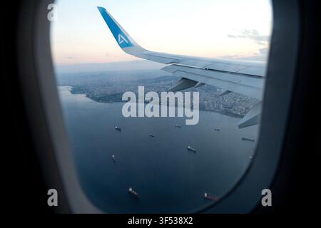 A view from an airplane window shows a wing extending over a city and water below. Stock Photo