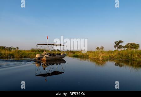 Safari boat reflected in a mirror-calm delta channel among papyrus reeds at dawn, Okavango Delta, Botswana Stock Photo