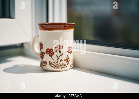 Close-up shot of a ceramic coffee mug featuring a hand painted red floral pattern resting on a bright white windowsill near a window with soft natural Stock Photo