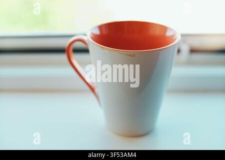 Close-up of an empty ceramic coffee mug featuring an orange interior and handle standing on a white surface near a window with soft light filtering th Stock Photo