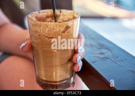 A close-up shot of a person holding a cold iced coffee beverage with thick foam in a transparent glass cup, resting on a wooden surface. Stock Photo