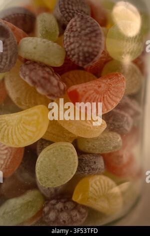 macro close-up of assorted frosted fruit-shaped hard candies piled in a glass jar; warm pastel yellows, oranges, greens and purples, soft shallow focu Stock Photo