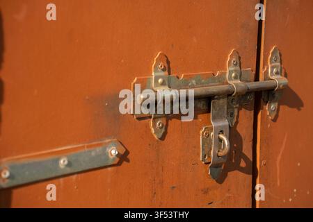 close-up of a weathered rusty metal latch and sliding bolt on an orange painted door, showing peeling paint, patina, screws and textured shadows in wa Stock Photo