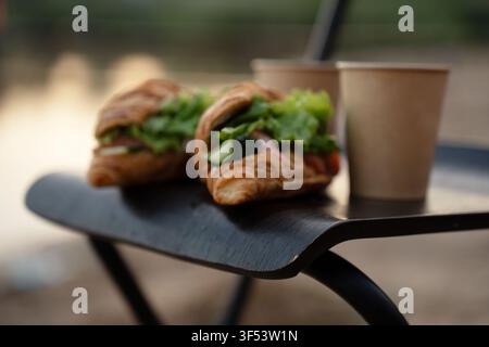 two croissant sandwiches stuffed with fresh lettuce, cucumber and tomato beside brown paper coffee cups on a curved dark outdoor table, warm morning l Stock Photo