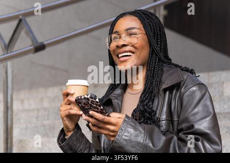 Smiling young woman using smartphone and drinking coffee outdoors Stock Photo