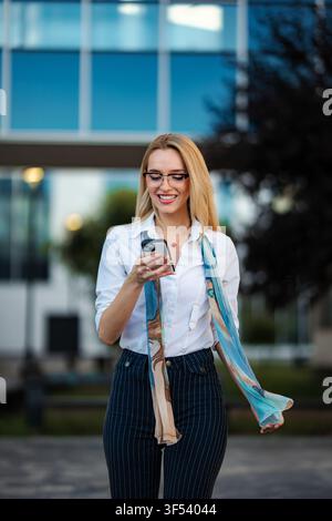 Happy businesswoman walking outdoors smiling using smartphone Stock Photo