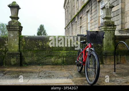Red bicycle with a black basket standing on wet cobblestones next to a moss covered stone wall, with an ancient stone building visible in the backgrou Stock Photo